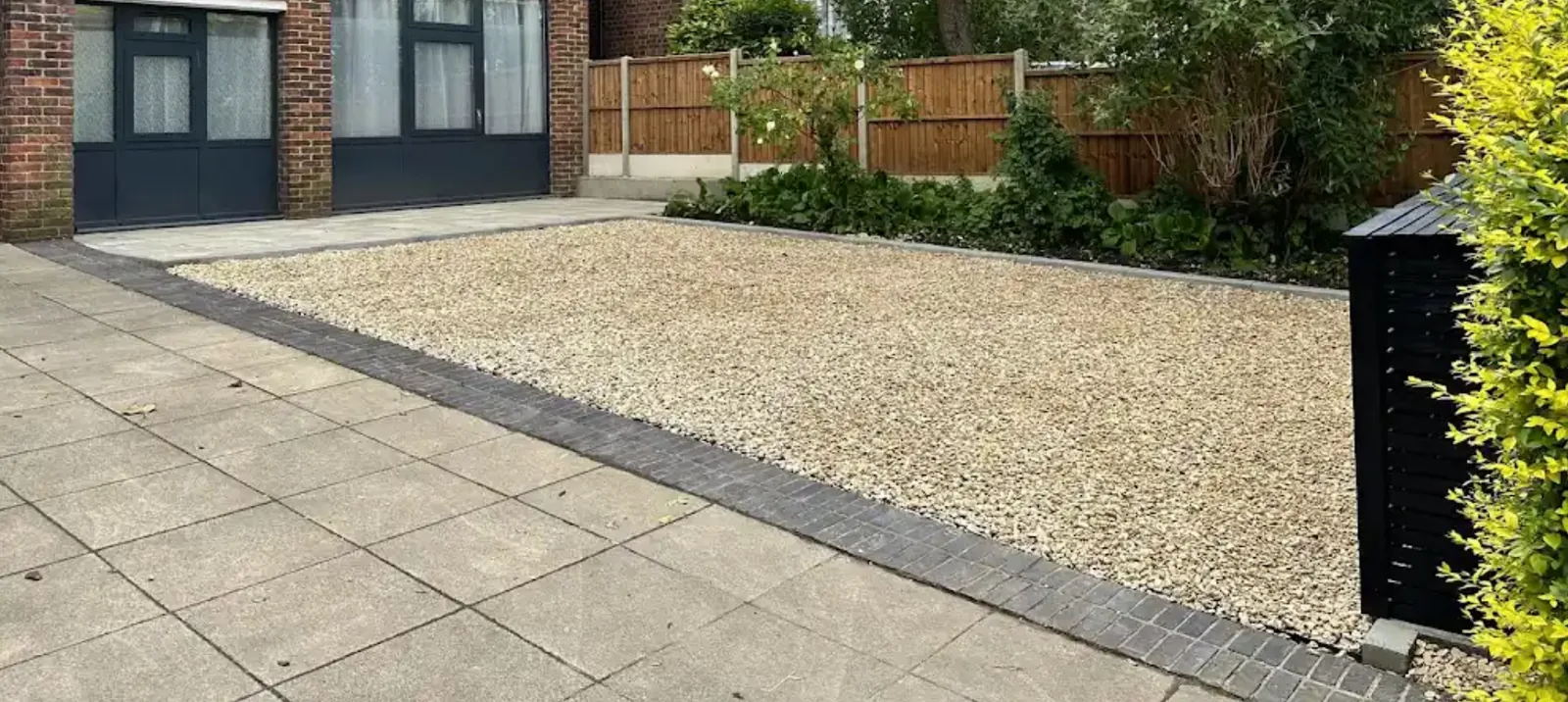 Gravel driveway with a paved border and block edging in front of a brick house with grey doors