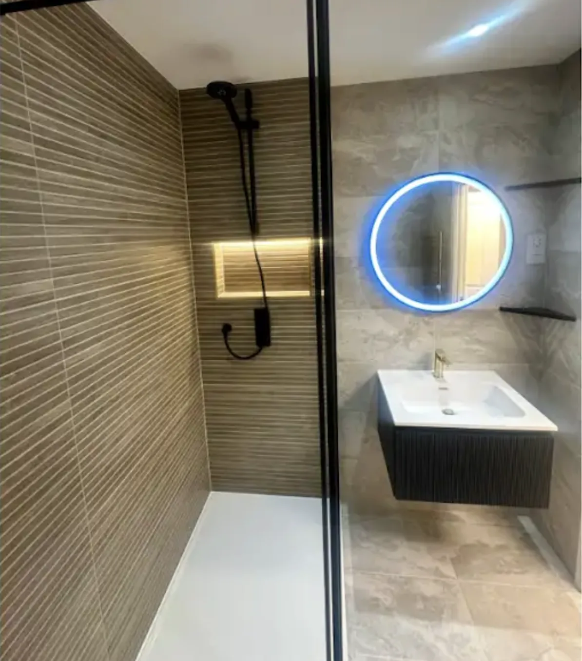 Walk-in wet room with stone effect wall tiling, a black framed shower screen, wall hung vanity and a round LED mirror